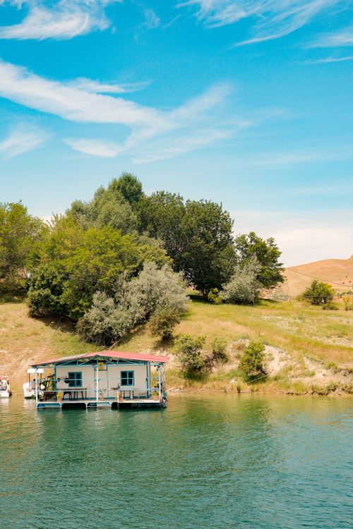 Free A charming houseboat by a vibrant riverbank surrounded by lush greenery under a clear blue sky. Stock Photo