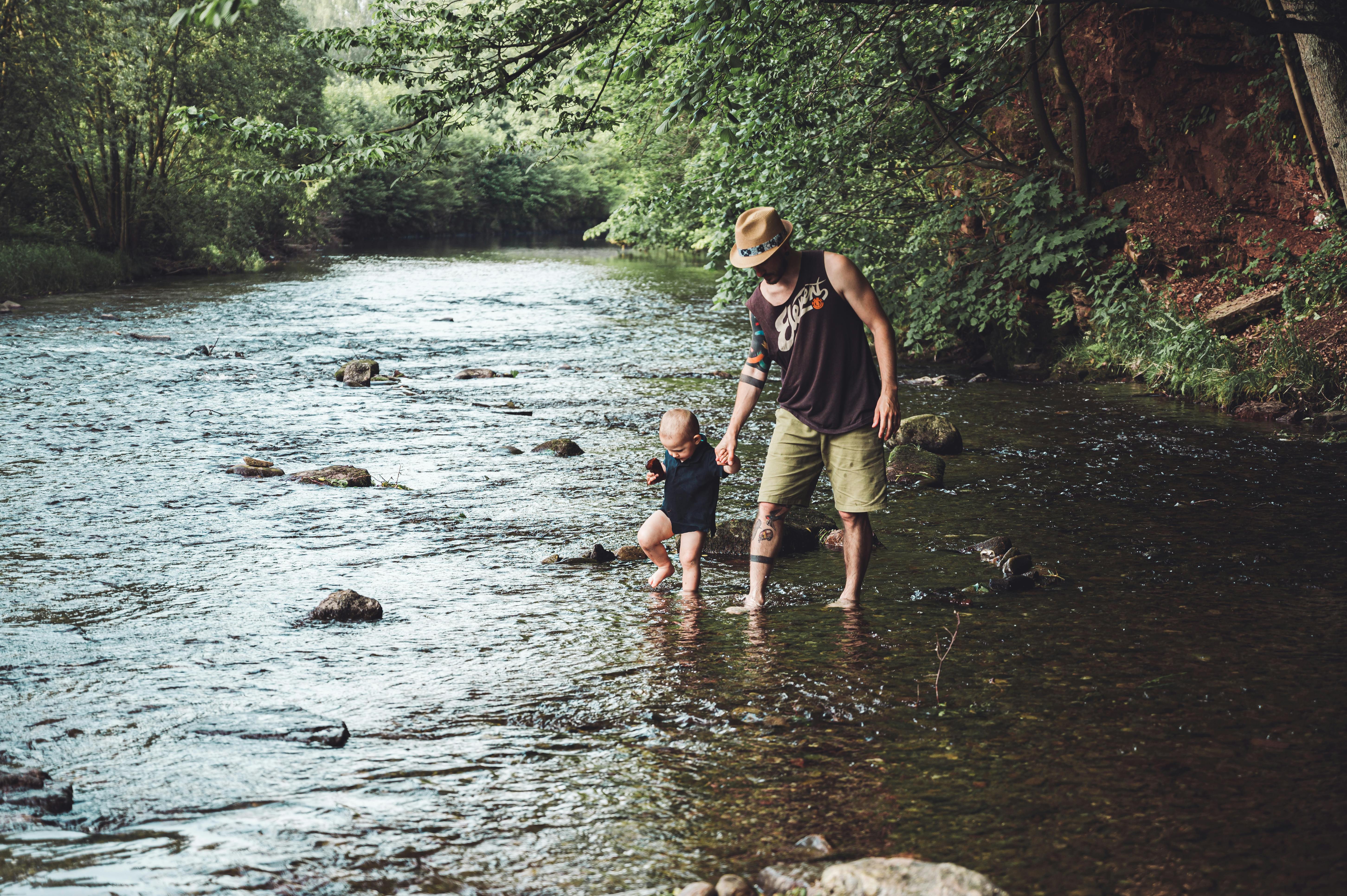 photo of man holding his child while walking in the river