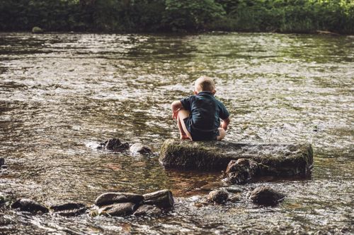 Free A young child sitting on a rock, enjoying nature in a tranquil river setting. Stock Photo