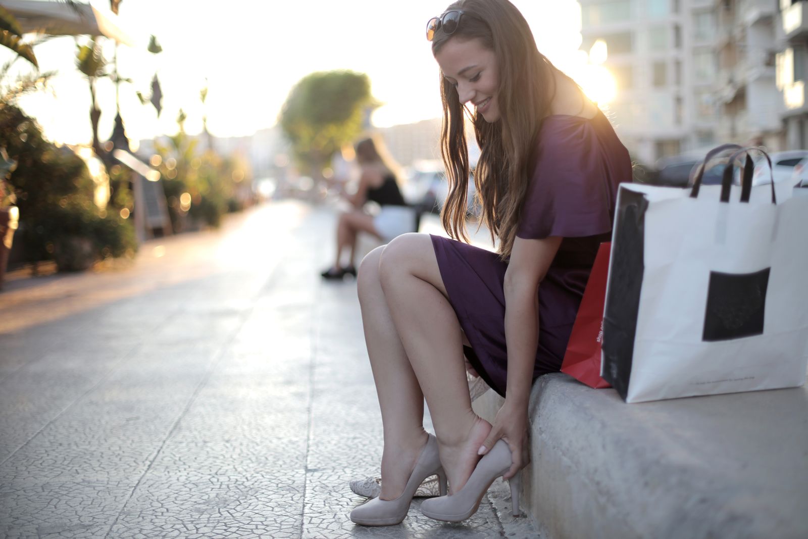 Smiling woman in a purple dress sits on a sunlit city ledge adjusting her high heels