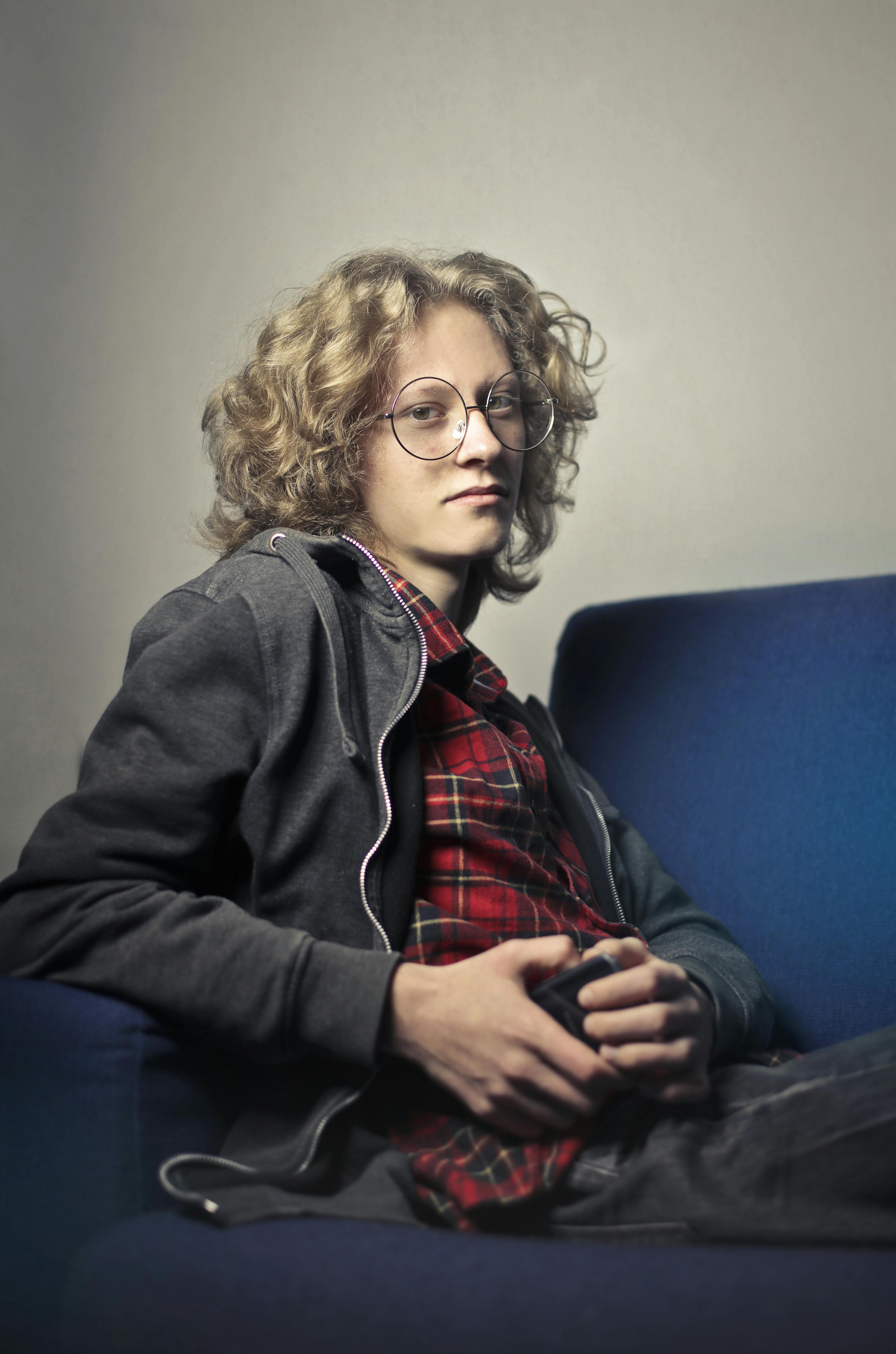 Free Pensive young man with eyeglasses and curly hair sitting on a blue sofa indoors. Stock Photo