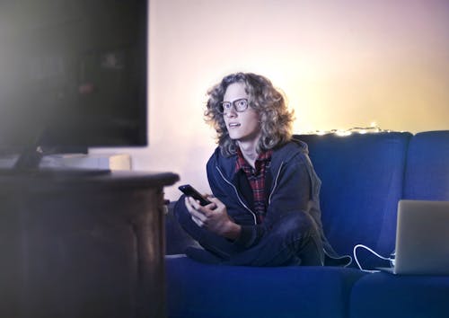 Concentrated male with long hair sitting on comfortable sofa at home and messaging on social media via cellphone while watching movie on TV with opened mouth