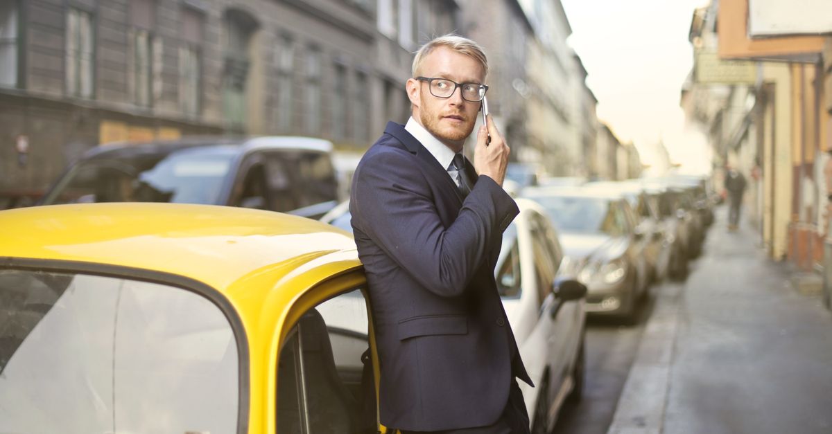 Businessman in a suit leaning on a yellow car making a phone call on a city street.