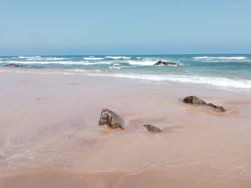 Free Quiet beach scene with gentle waves and exposed rocks under a clear blue sky. Perfect for relaxation themes. Stock Photo