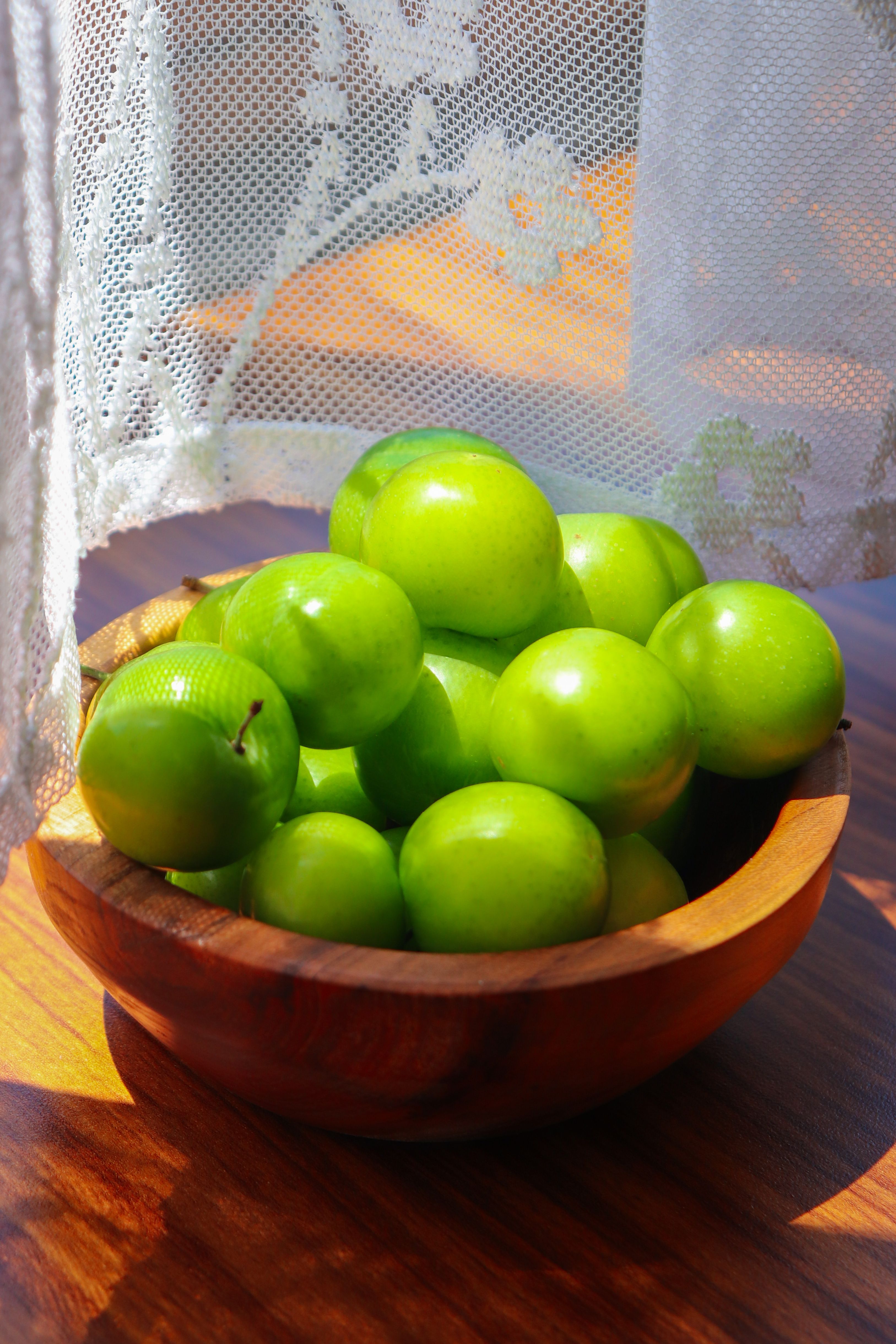 A bowl of fresh green plums in sunlight, placed near a lacy white curtain.