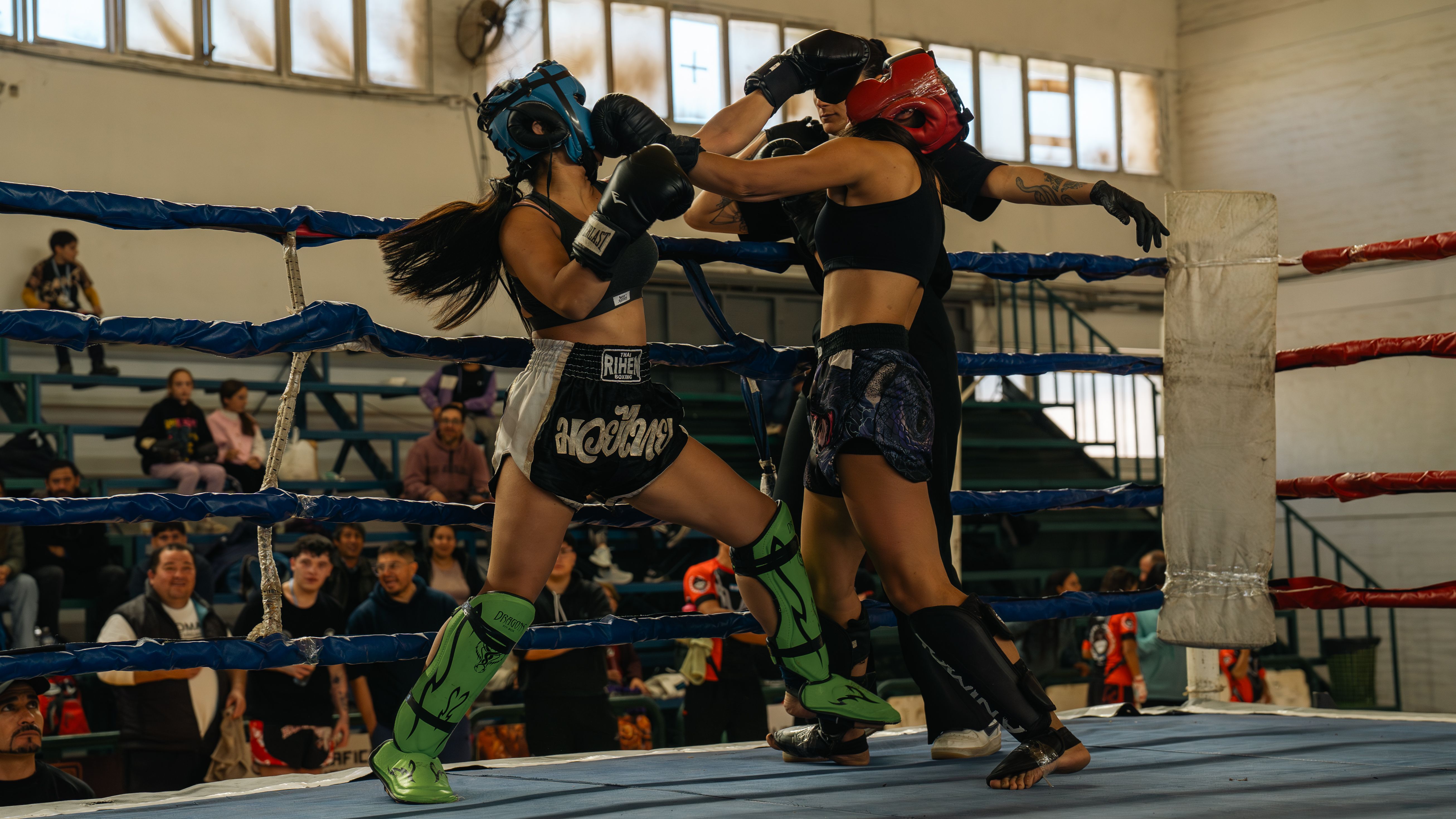Two women competing fiercely in a Muay Thai boxing match inside a gym with an audience watching.