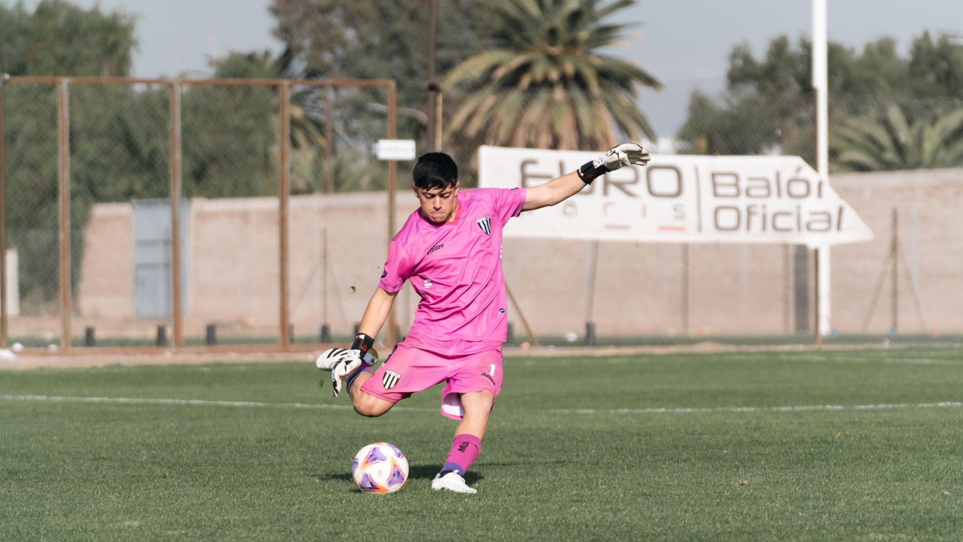 Goalkeeper in pink uniform making a kick during a soccer match outdoors.