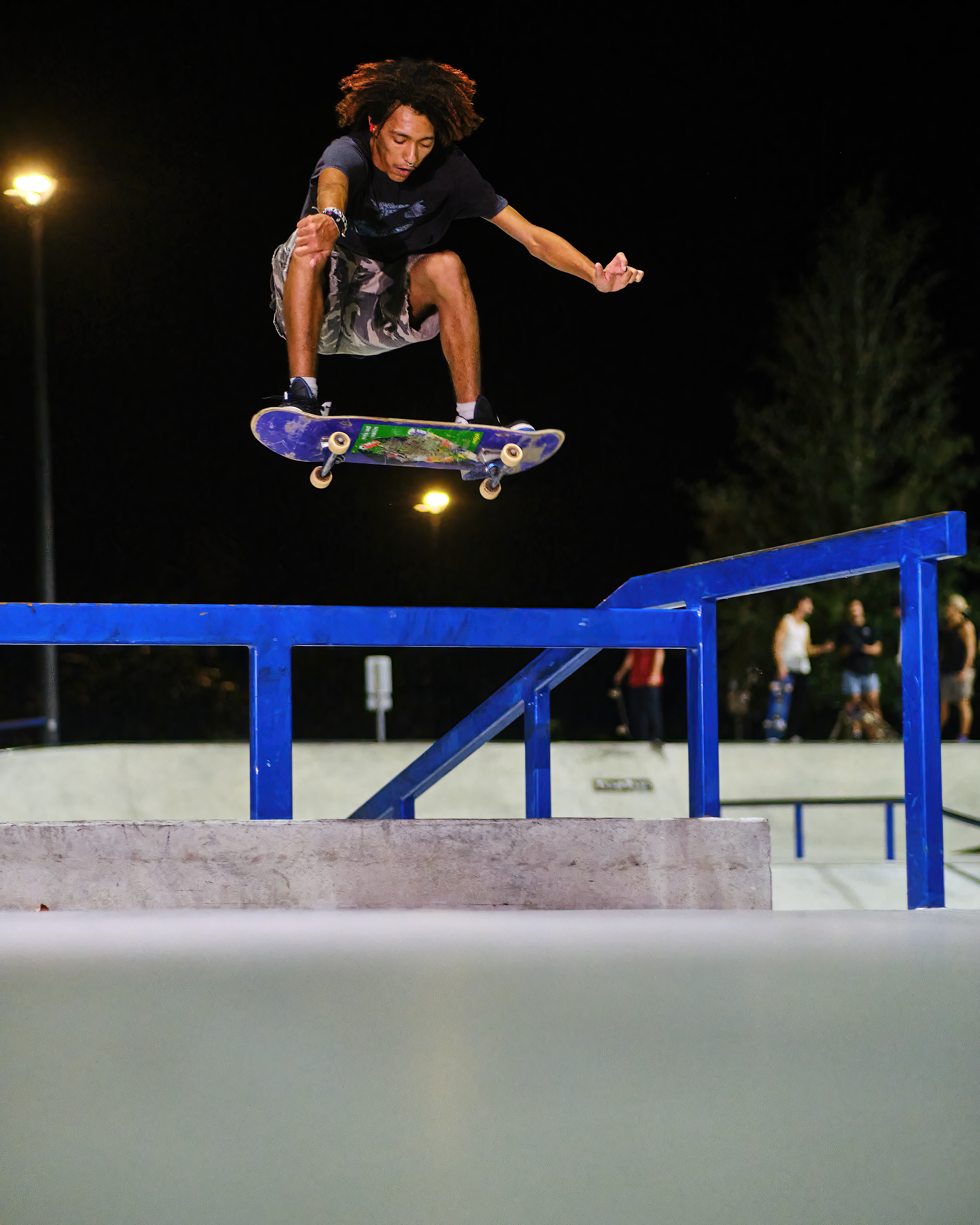 Skateboarder executing a jump trick over a rail at a skatepark at night.