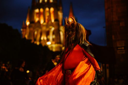 A vibrant costume parade participant stands in front of an illuminated cathedral at night.