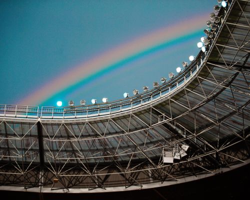Free Bright rainbow arches over a modern stadium's metal roof, creating a vivid contrast against the sky. Stock Photo