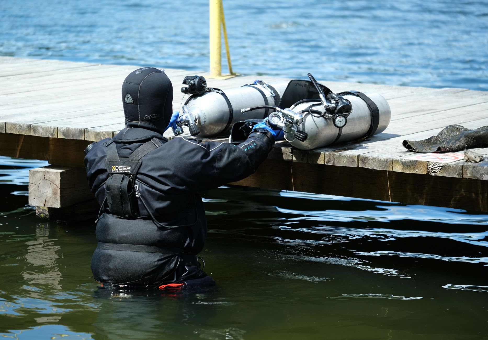 A scuba diver in a wetsuit prepares diving equipment on a lakeside wooden dock.
