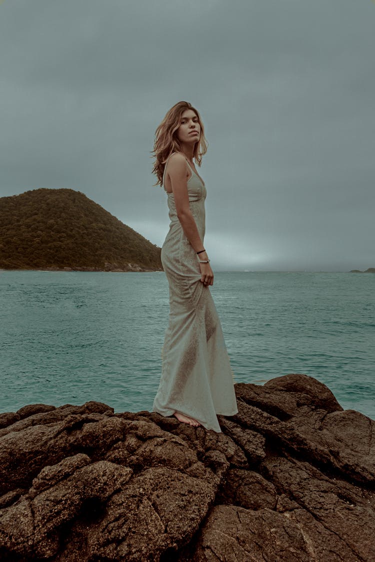 Woman In White Sleeveless Dress Standing On Brown Rock Near Body Of Water