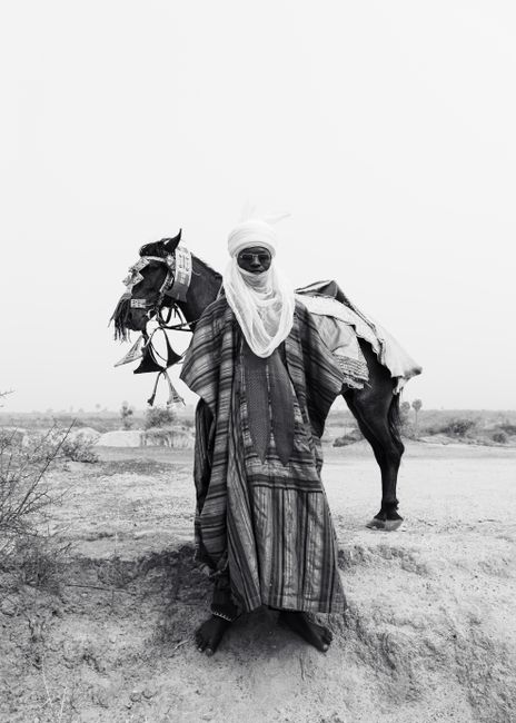 A nomadic man in traditional attire stands beside a horse in a desert landscape.