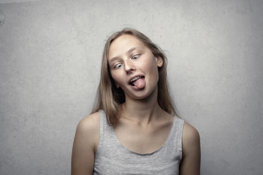 A young woman with a playful expression sticks out her tongue against a gray background.