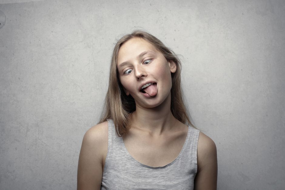 A young woman with a playful expression sticks out her tongue against a gray background.