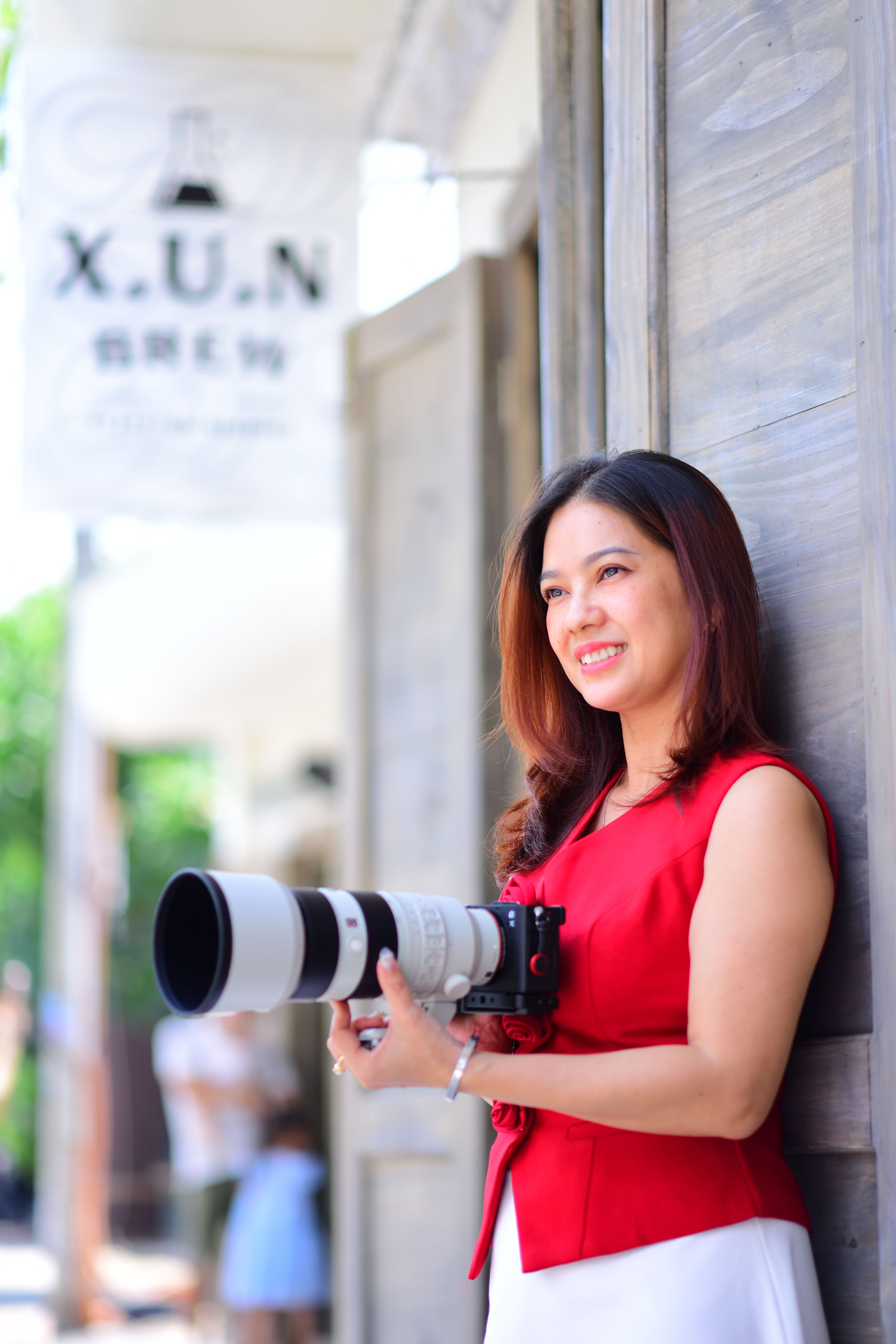 A cheerful woman holding a camera with a large lens in an urban outdoor setting.