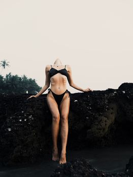 A woman in a bikini leans back on rocky terrain at the beach.