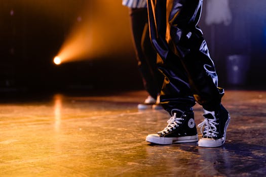 舞台上，一名穿着黑色运动鞋和闪亮裤子的舞者被明亮的灯光照亮，特写镜头聚焦在他身上