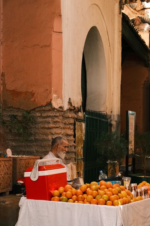 Free A street vendor stands beside a cart of ripe oranges under an archway in an outdoor market. Stock Photo
