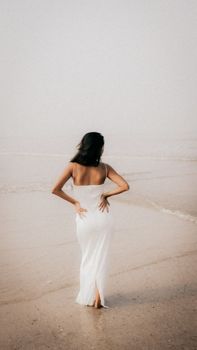 Elegant woman in a white dress walking on a foggy beach, exuding calm and tranquility.