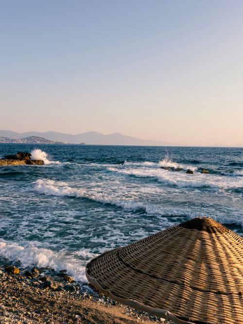 Free Scenic sunset view of Aegean Sea waves crashing with a straw beach umbrella in the foreground. Stock Photo