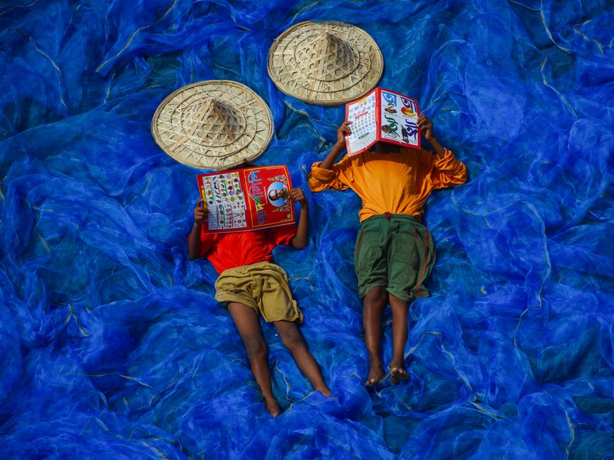 Two children reading books on blue fishing nets in West Bengal, India, wearing traditional hats.