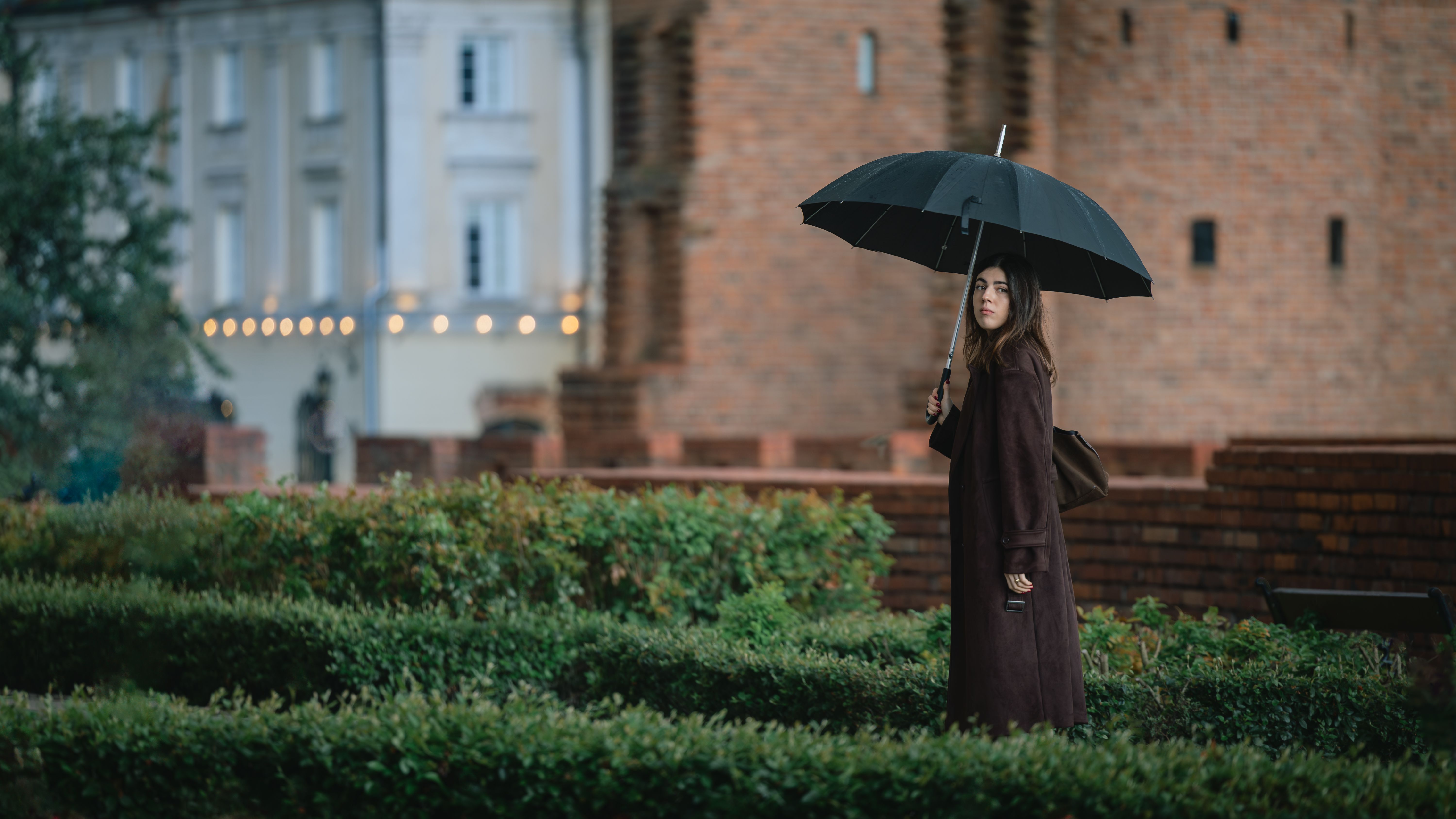 A woman holding an umbrella stands near historical brick architecture during a cloudy day.