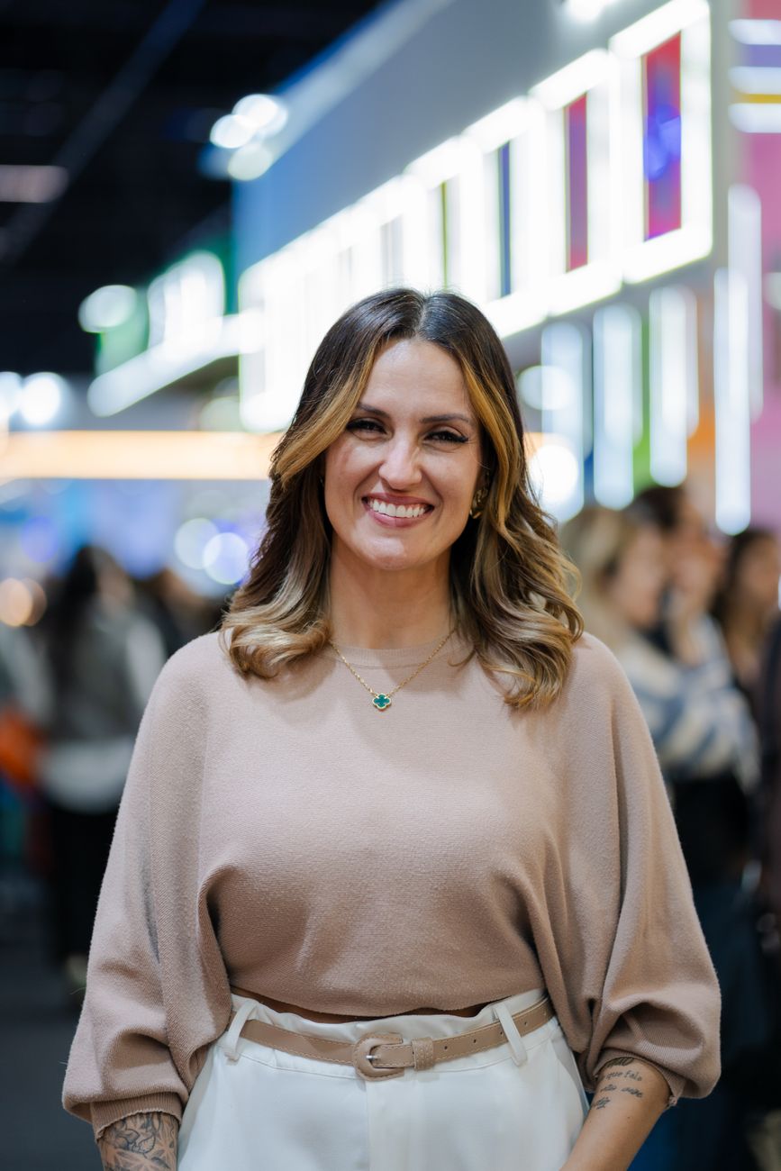 Retrato de uma mulher sorridente em um evento interno com luzes coloridas desfocadas e uma multidão ao fundo.