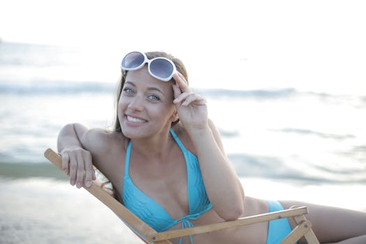 A woman in a blue swimsuit enjoys a sunny day at the beach, reclining on a folding chair with sunglasses.