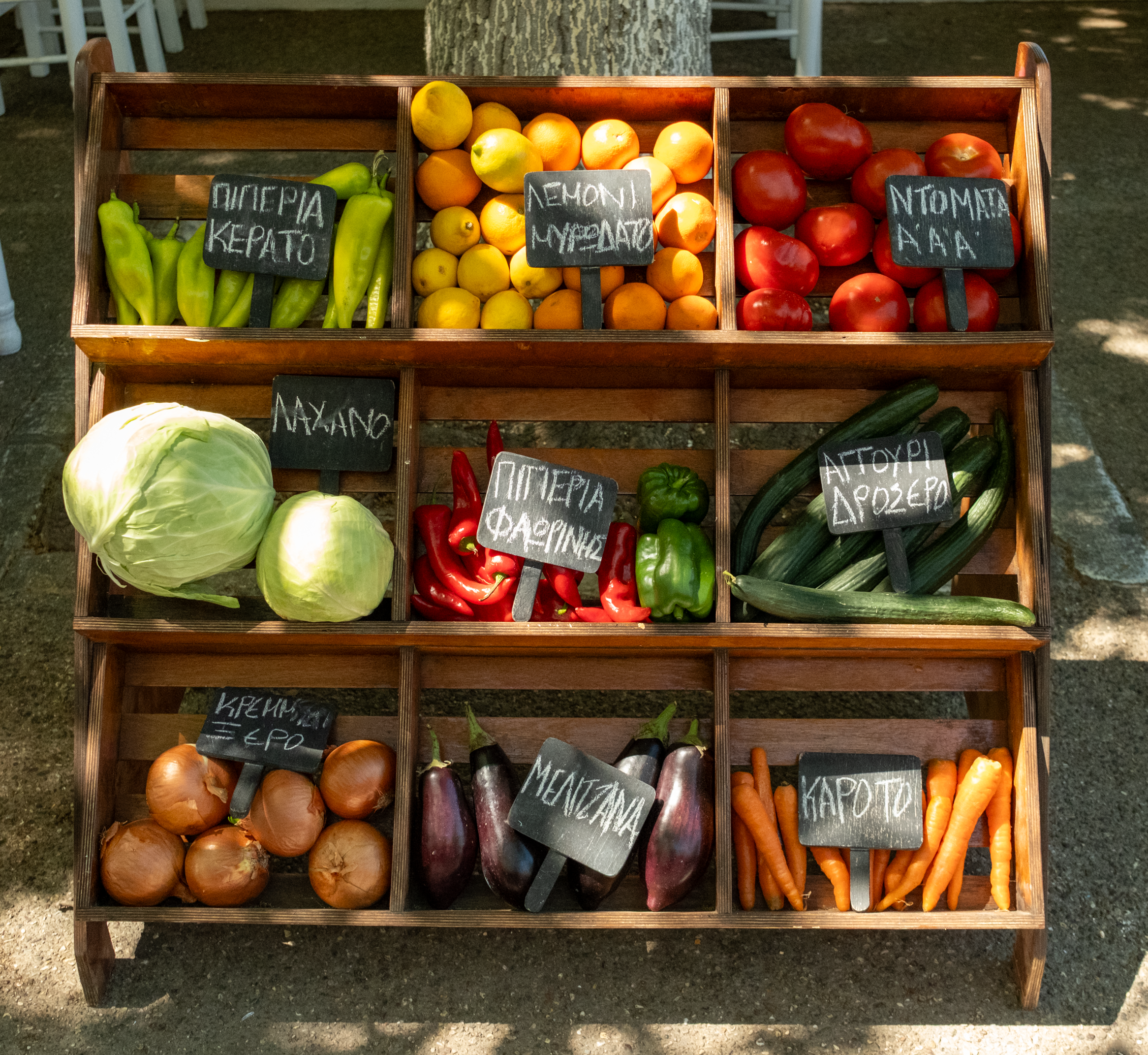 Wooden shelf displaying fresh vegetables and fruits at market with Greek labels.