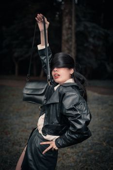 Stylish woman posing in black leather jacket holding handbag against an outdoor backdrop.