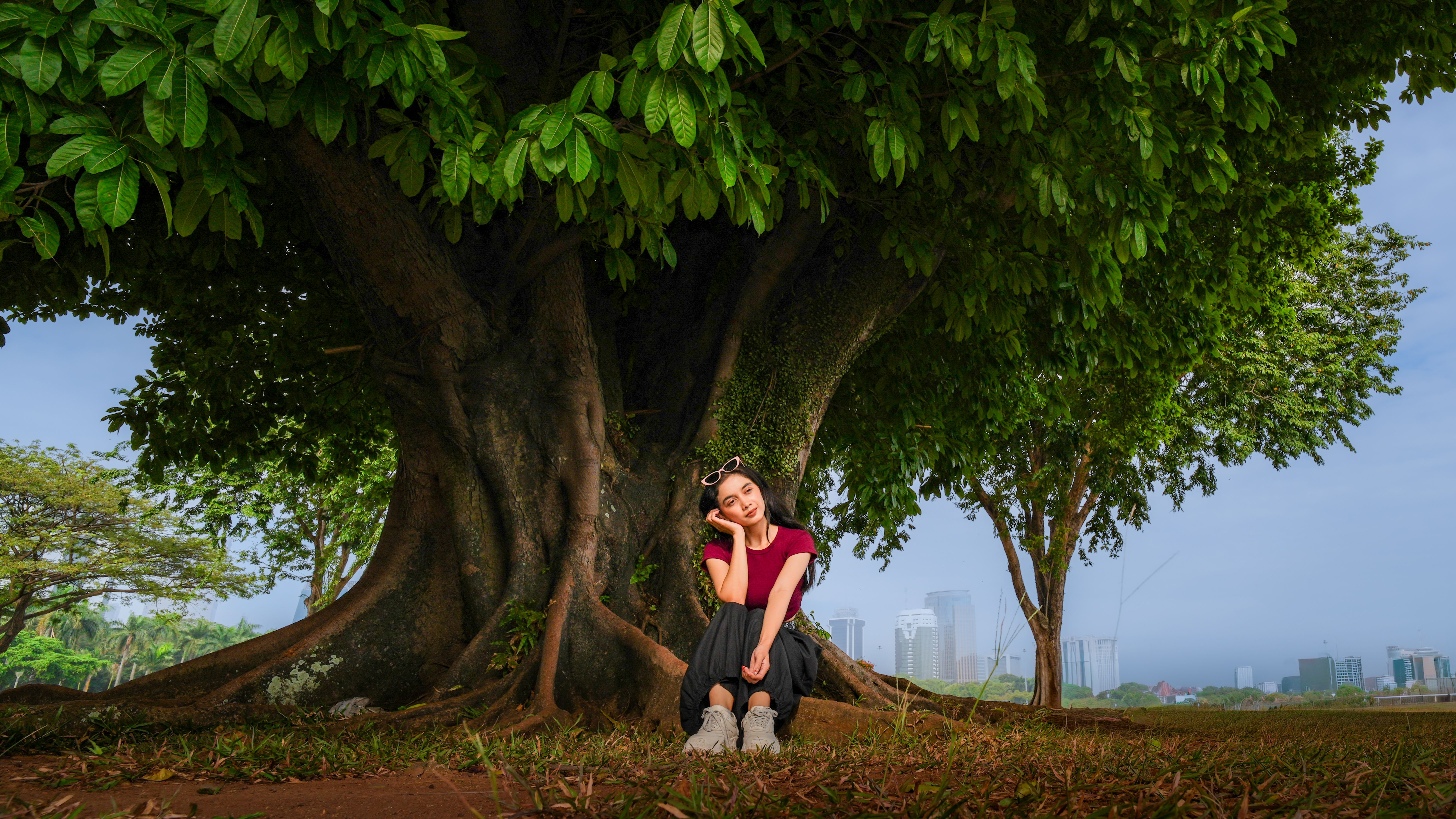 A young woman sits peacefully under a large tree in a Jakarta park, skyscrapers in the background.