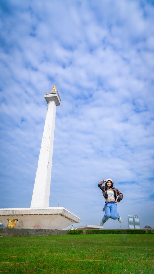 Free A woman joyfully jumps near the National Monument in Jakarta under a bright, cloudy sky. Stock Photo