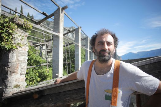Man standing outdoors in rustic setting, wearing suspenders and smiling in sunlight.