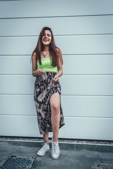 一位自信的年轻女子身着花卉裙子和霓虹灯上衣，在户外靠着白墙微笑