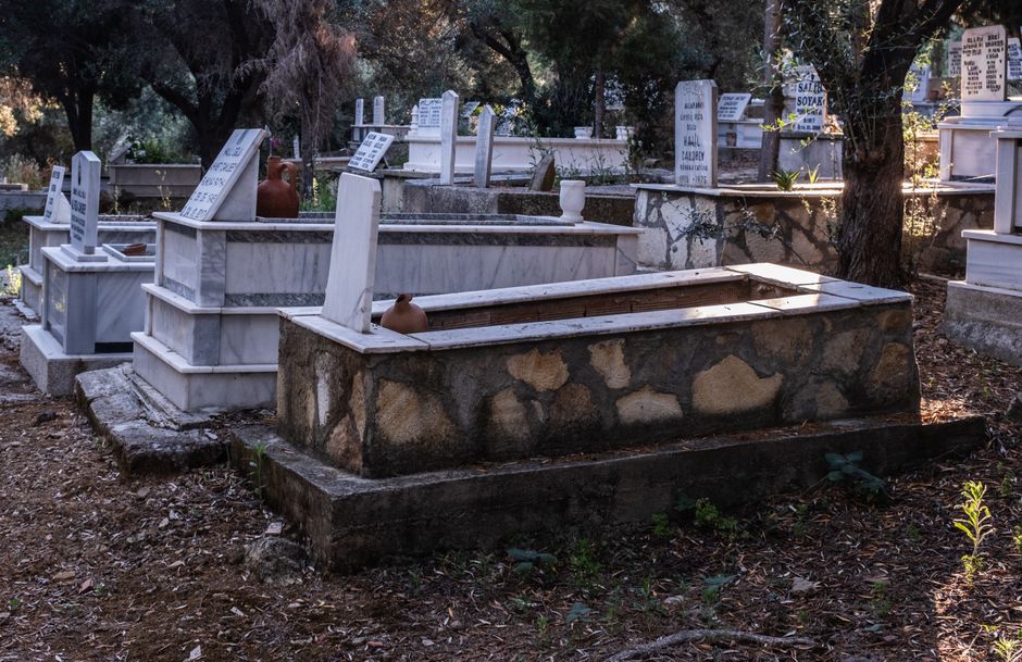 Fotomural Cementerio al aire libre pacífico en Foça, İzmir, con luz solar filtrándose a través de los árboles y lápidas de piedra.