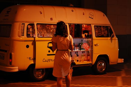 A woman at a vintage coffee van at night in Kaliningrad. Warm lighting sets a cozy urban scene.