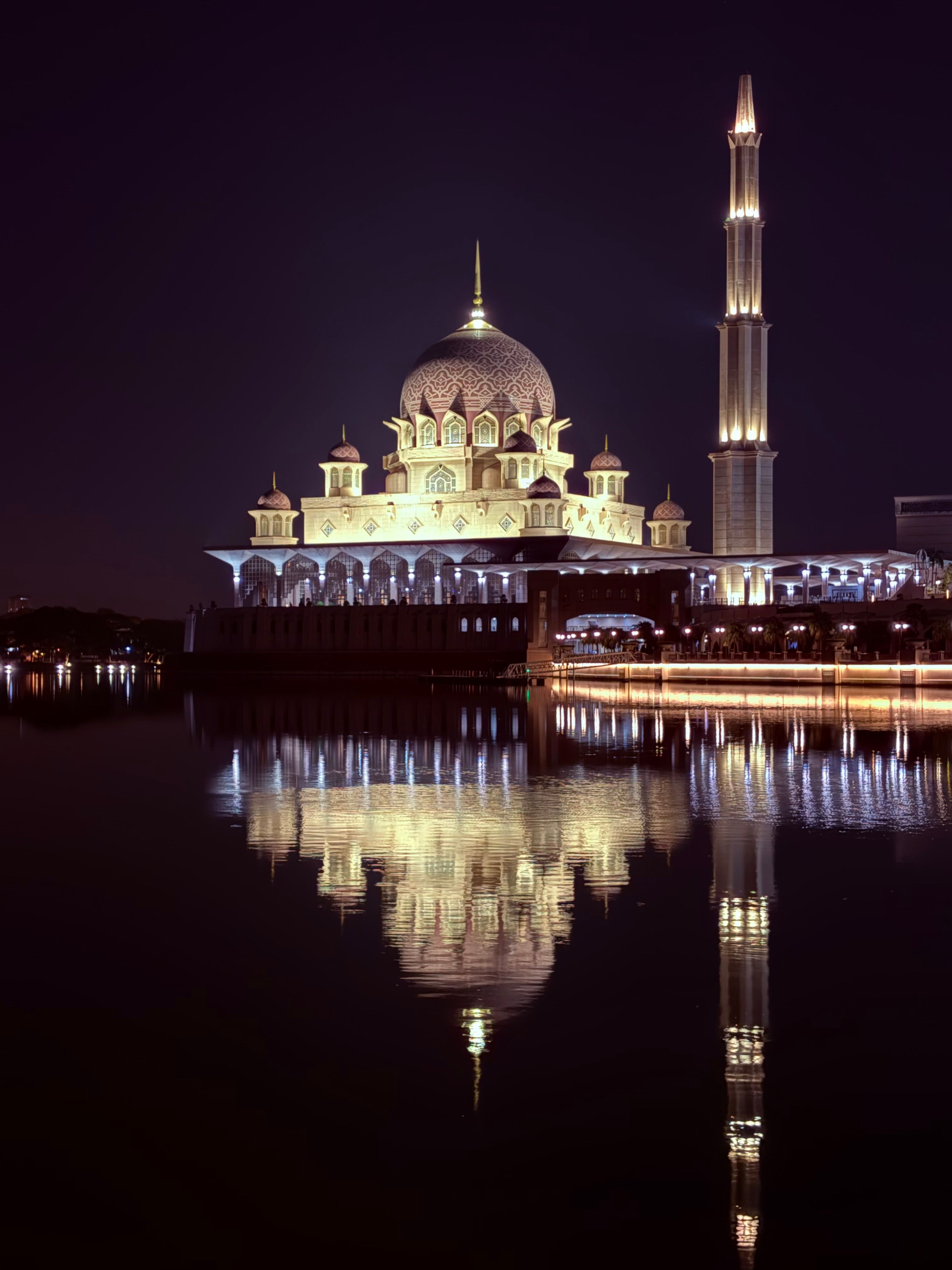 A beautifully lit mosque and minaret reflecting on water under the night sky in Malaysia.
