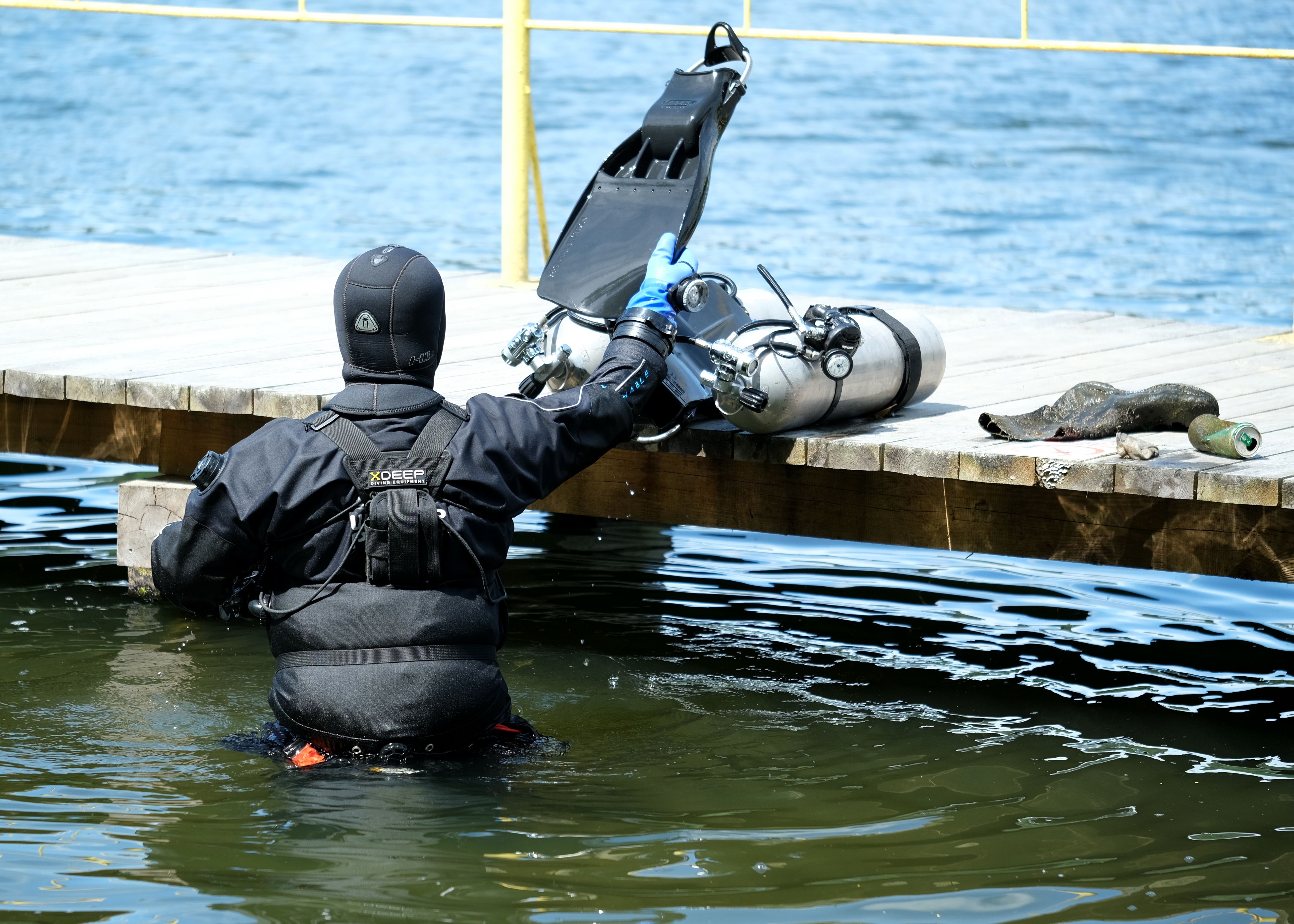 Scuba diver in wetsuit prepares scuba gear on a sunny pier by the water. Calm and outdoors setting.