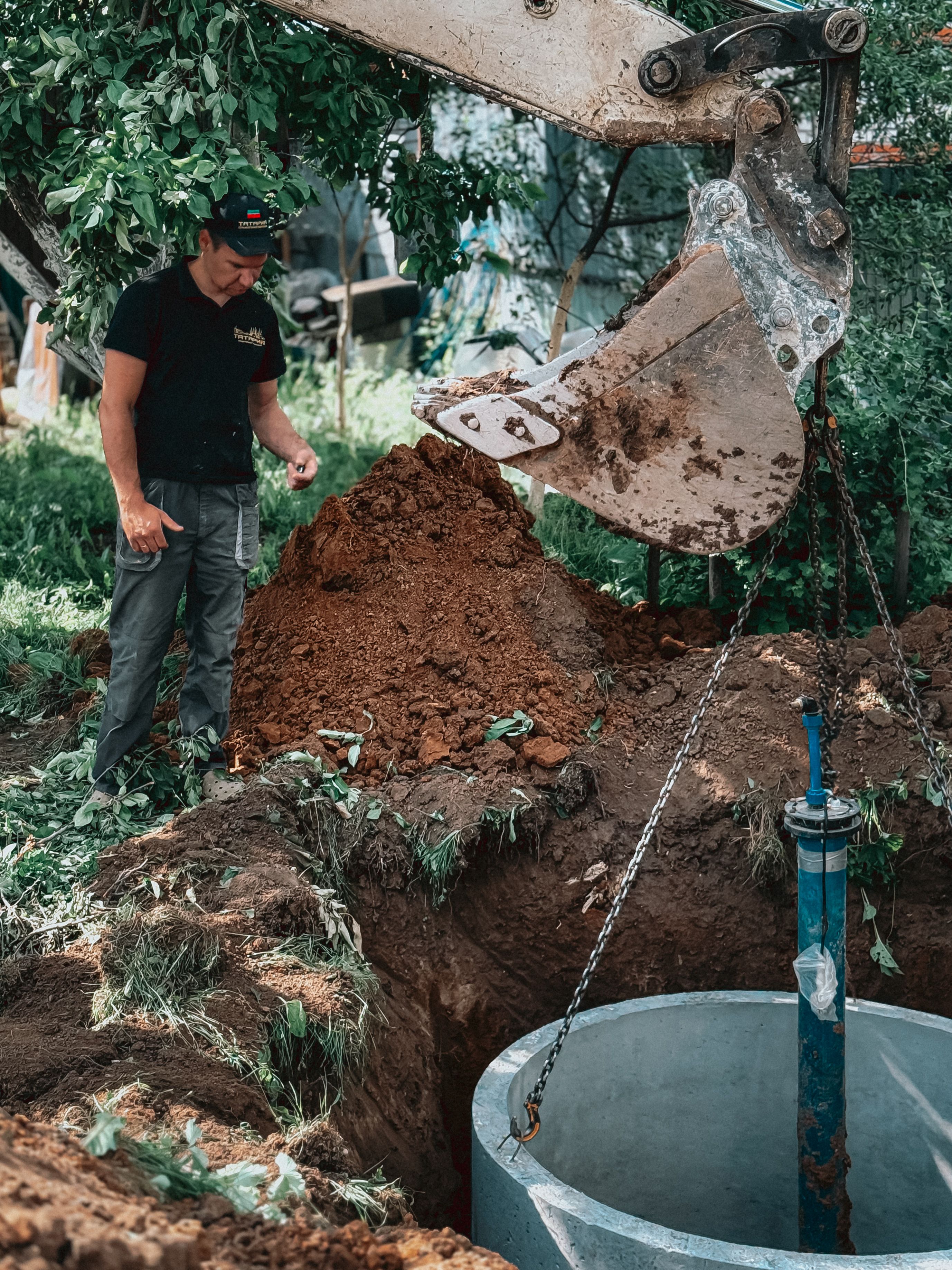 A worker supervises a concrete pipe being lowered with heavy machinery in a backyard.