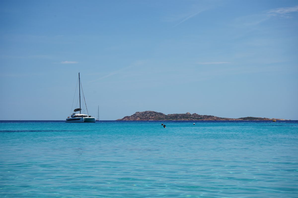 Catamaran ancorat in apa turcoaz laptoasa, cu o insulita pe orizont