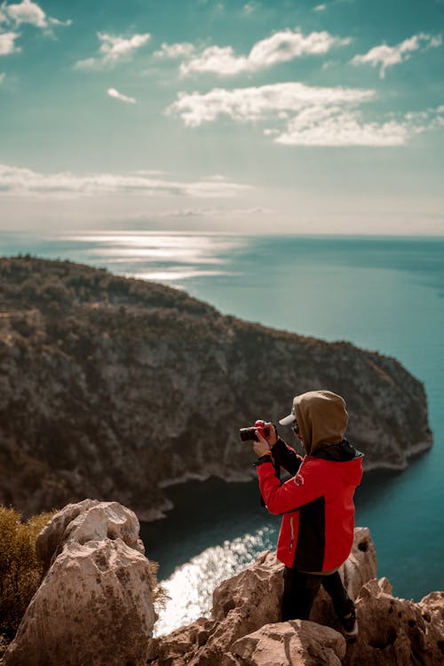 Man in Red Jacket Taking a Picture while Standing on Rock