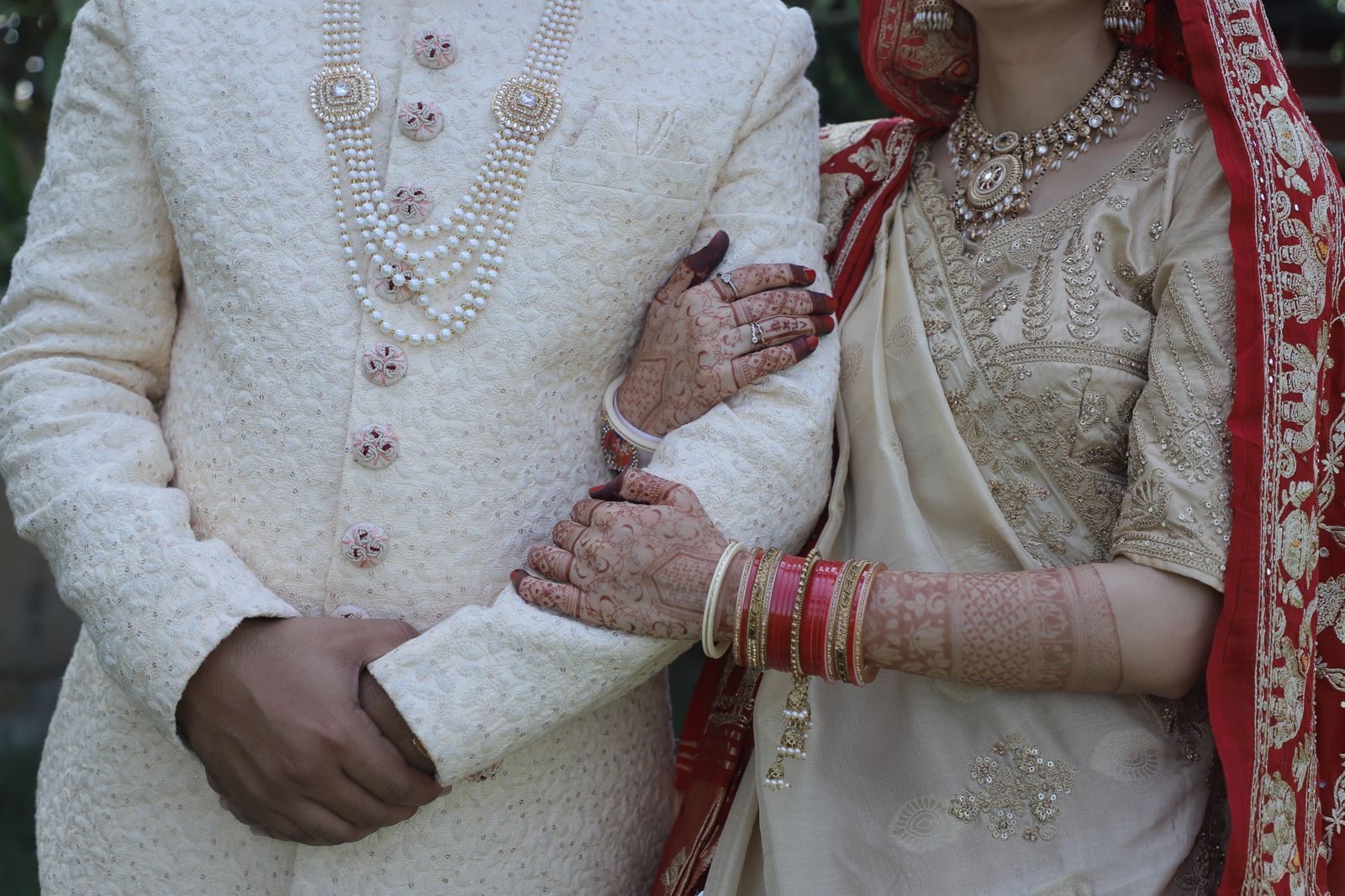 An ivory sherwani beside a pair of mehndi covered hands resting on a forearm, both heads out of frame.