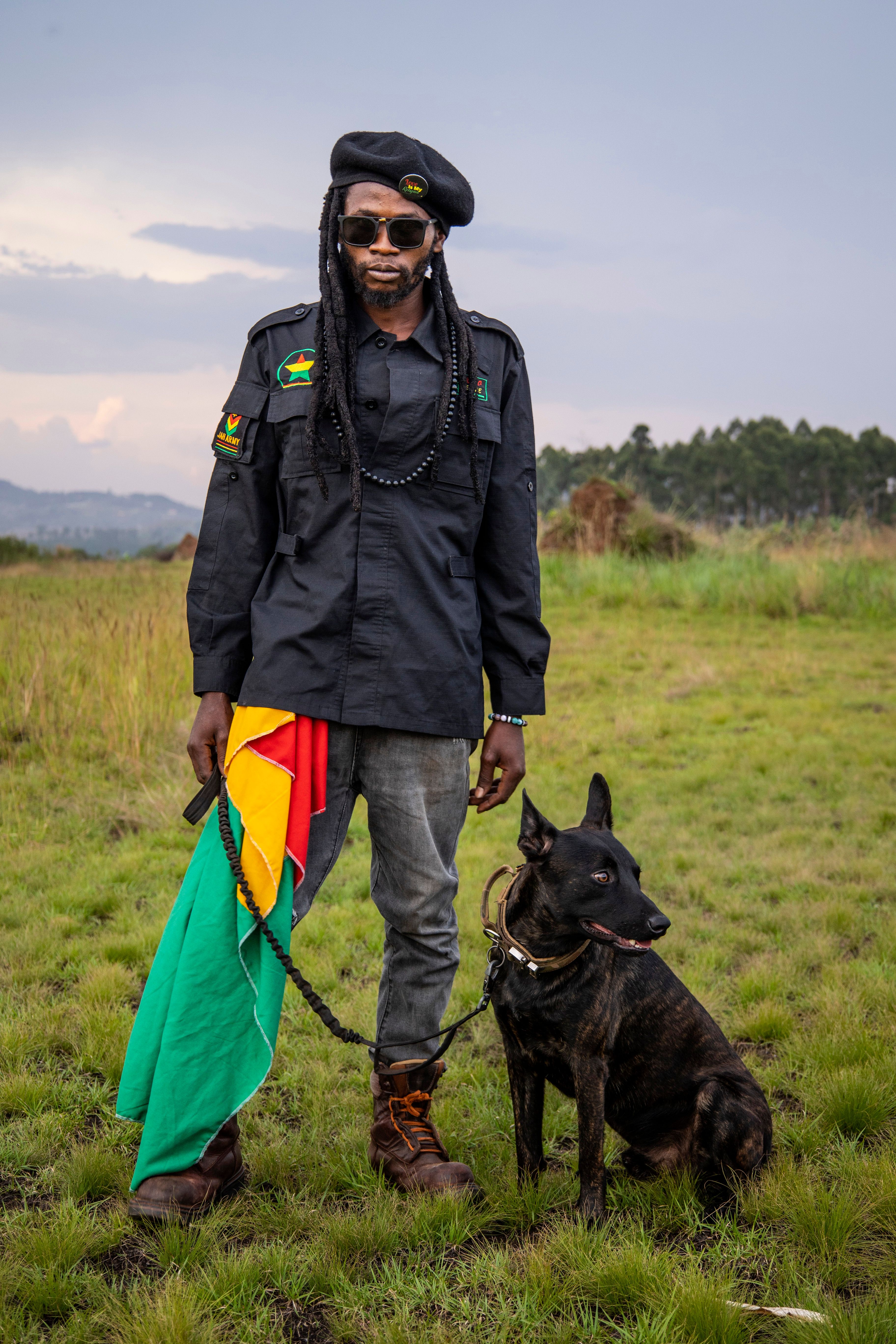 Man in military-style attire holds a dog on grassy field, with vibrant flag, outdoors in Uganda.