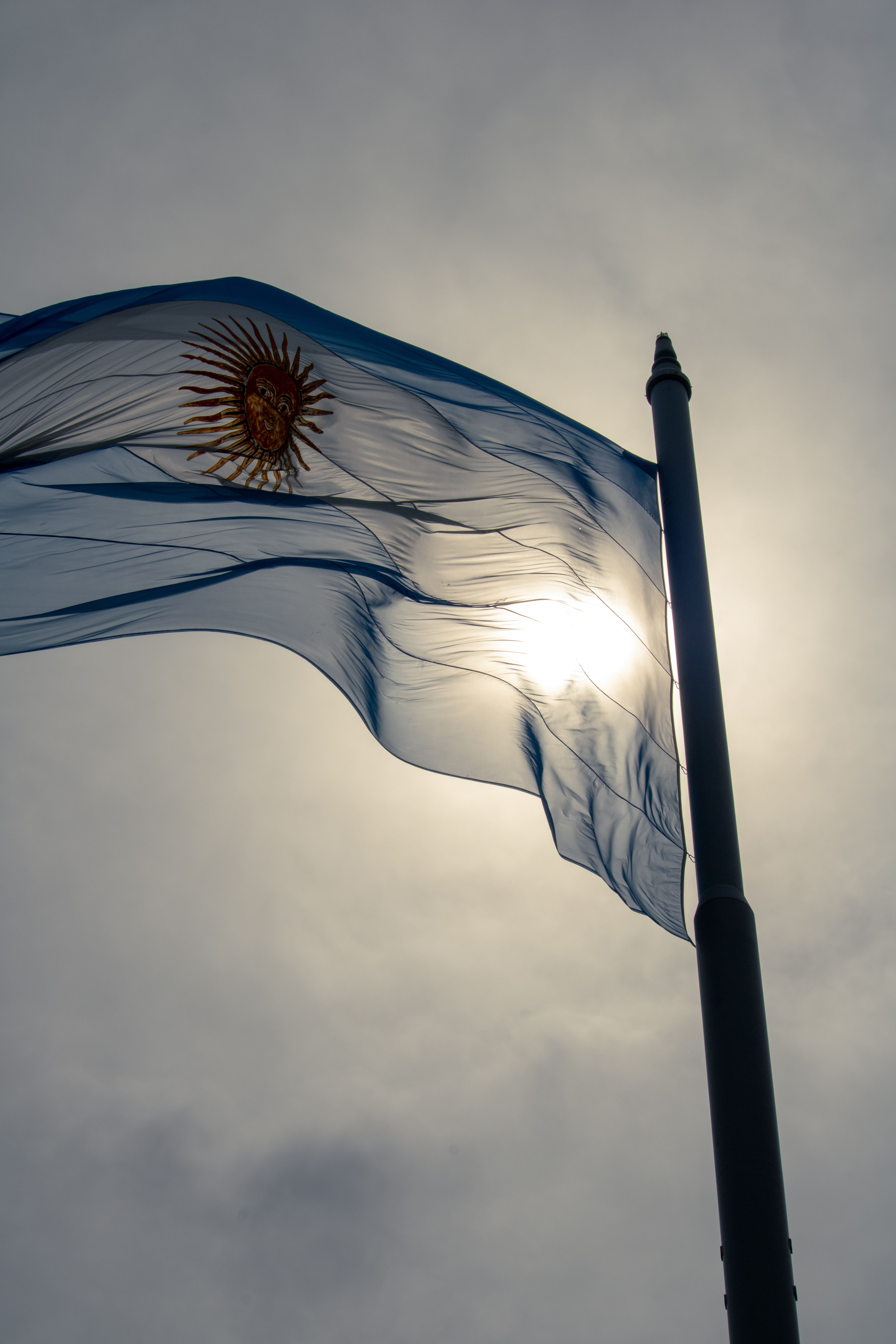Dramatic view of the Argentinian flag backlit by the sun against a cloudy sky, symbolizing national pride.
