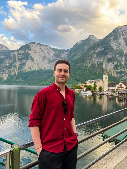 Free A man in a red shirt posing on a bridge with a picturesque lake and mountain backdrop in Austria. Stock Photo