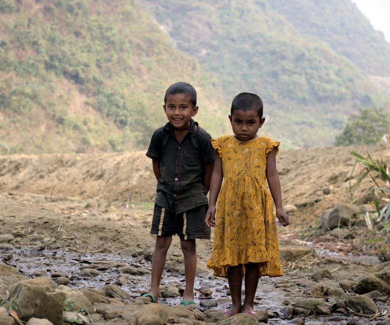 Two children smiling and standing on a rocky path amidst lush green hills on a sunny day.