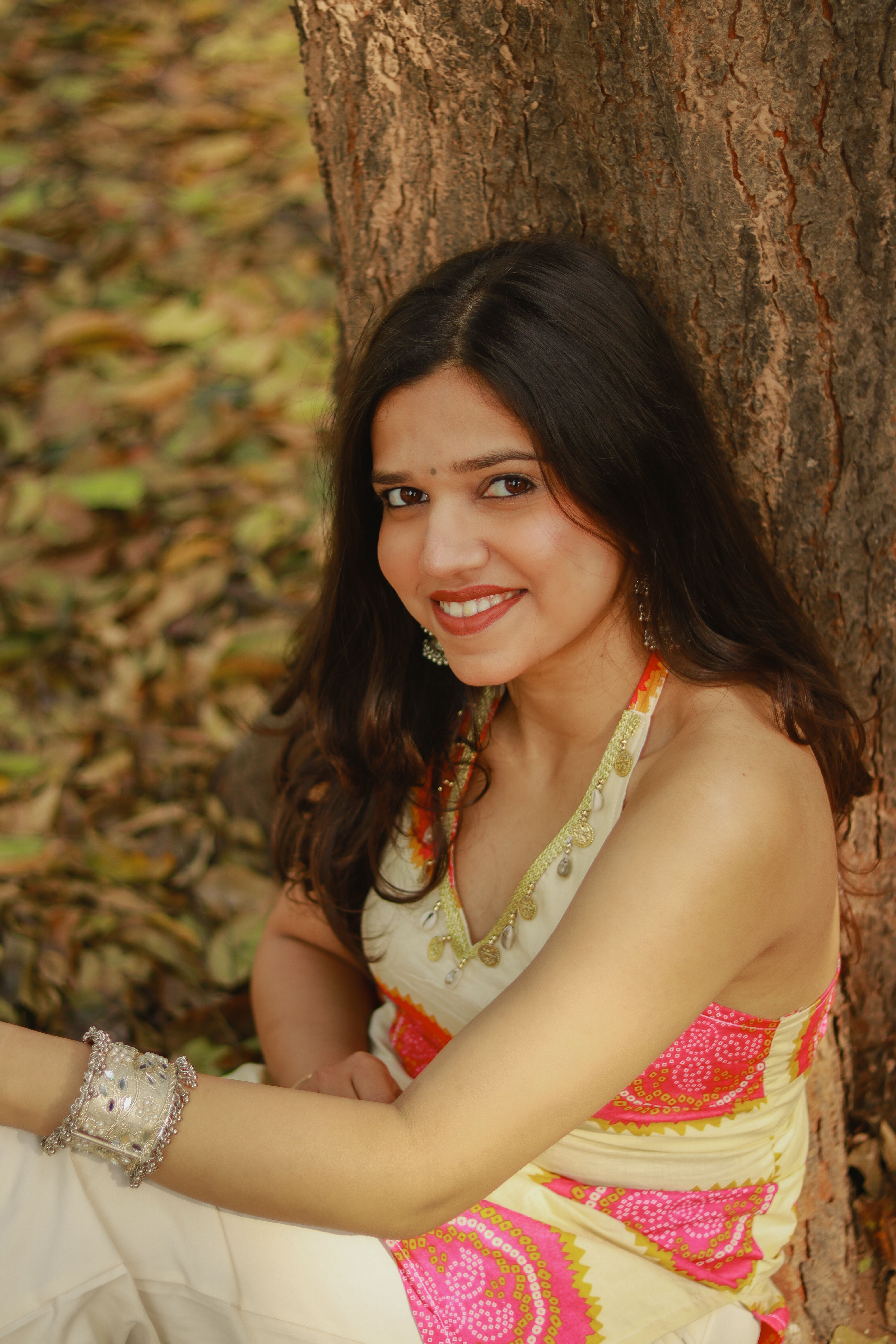 A cheerful woman in a colorful dress leaning against a tree in a natural setting.