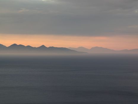 Calm sea at sunset with distant mountains under a soft, muted sky.