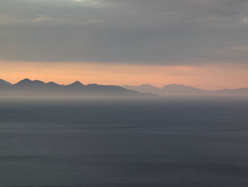 Calm sea at sunset with distant mountains under a soft, muted sky.